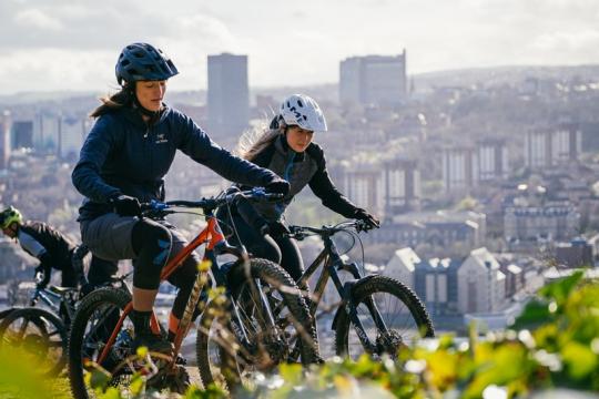 Two cyclists are on a hill overlooking Sheffield with the city in the back ground