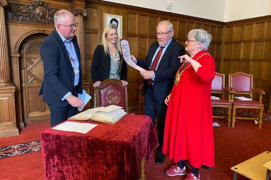 Two women and two men stand in one of the Town Hall rooms, over a table with a book and red table cloth on it