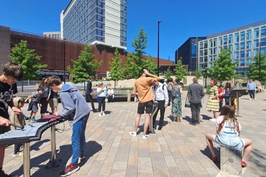 Several people can be seen playing with musical instruments with a road in the middle ground and buildings in the back ground