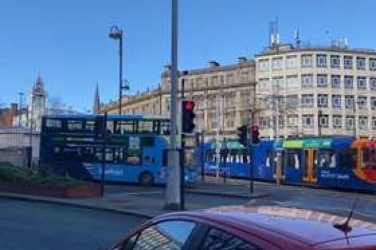 A picture of Sheffield city centre with a bus and a tram crossing over the roads with a car in the foreground and buildings in the back ground