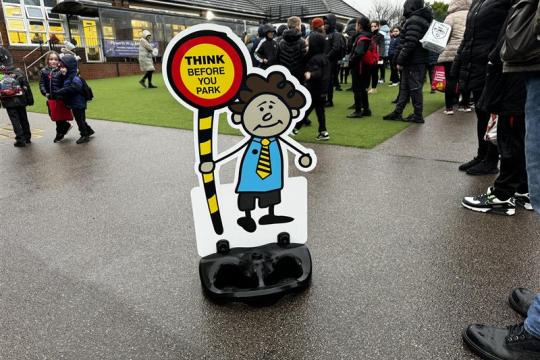 A cut out of a cartoon child holding a road crossing lollipop stands in a playground surrounded by children in their school uniform