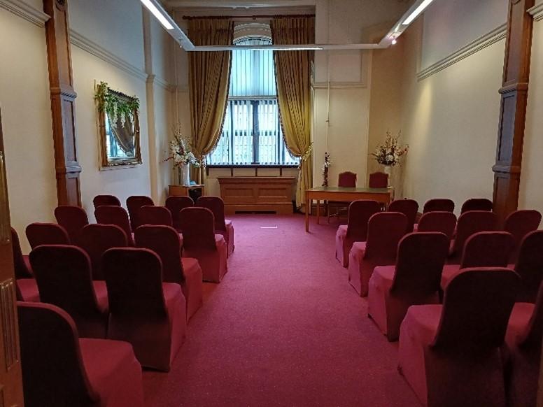 Two rows of seats dressed in red with a central aisle face a desk positioned to the right with two chairs. A small plinth with a large bouquet of flowers is positioned to the right. Behind all of these is a window with a blind and golden curtains that are tied back. A mirror decorated with a flower garland is visible on the left hand wall.