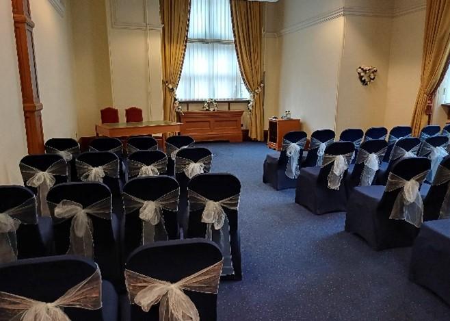 Two rows of navy blue seats with white ribbons tied at the back face the front with a central aisle between them. A table with two chairs is placed to the left at the top of the room facing the rows. Behind the table and to the right are windows with blinds and gold-coloured curtains tied back.