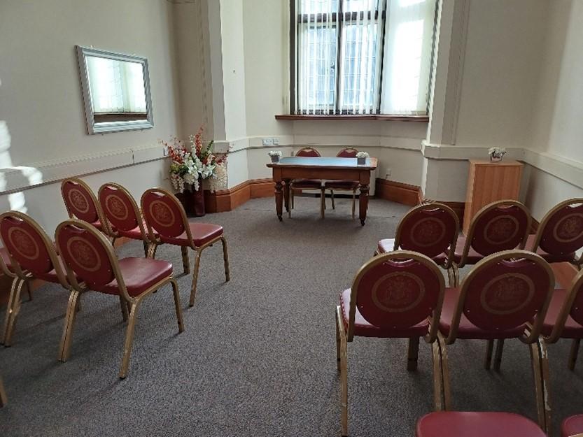 Rows of seats with a central aisle face a table with chairs behind. The table is placed in front of a long window with a blind. A small table stands to the right of the central table and a large bouquet of flowers sits to the left. A mirror is visible on the left wall.