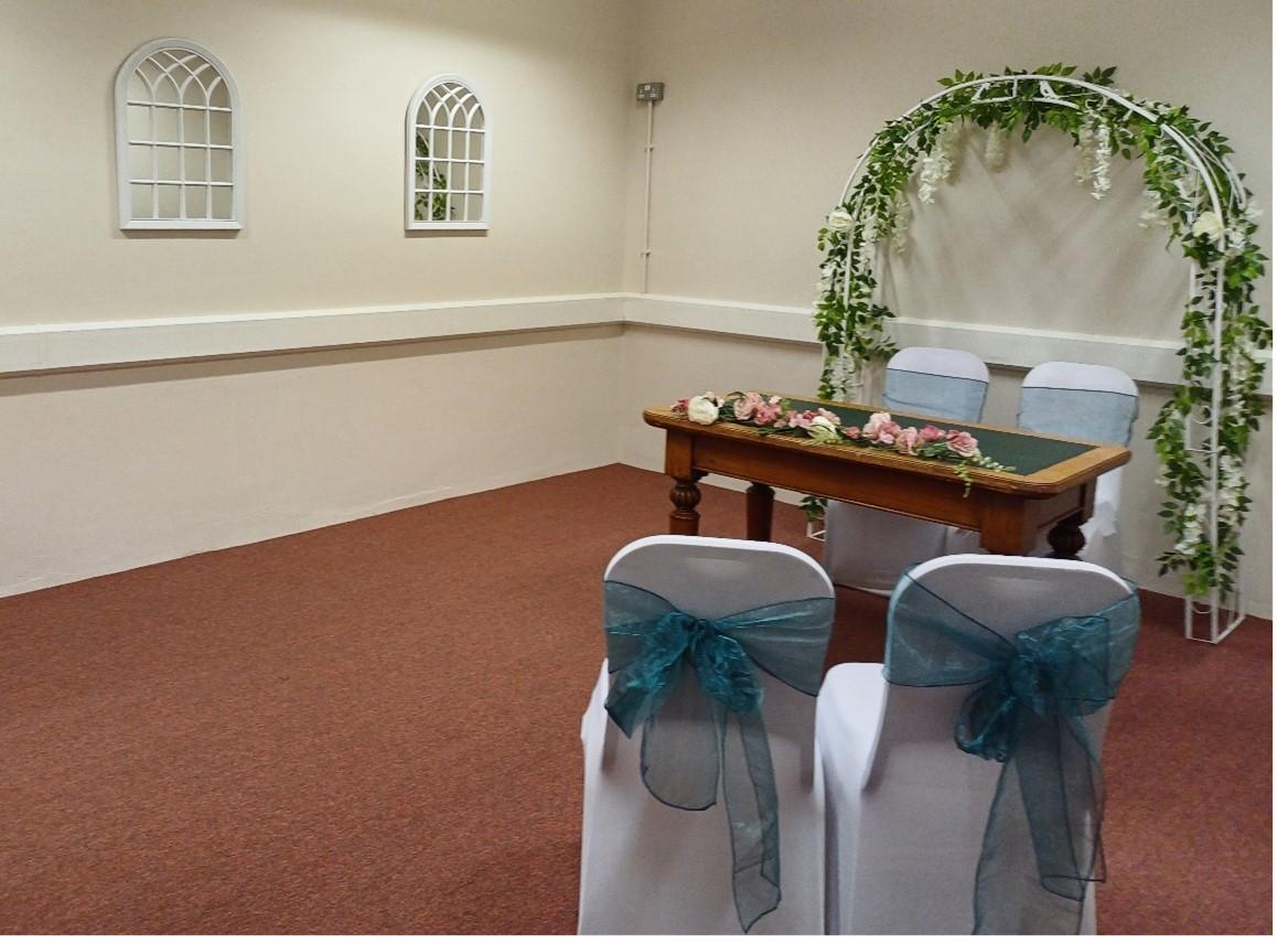 A table with two chairs and a flower arch behind it stand against the wall, facing you as you enter the room. Two small white mullioned internal windows are positioned on the left hand wall and two dressed white chairs with blue bows tied at the back are positioned slightly to the right and facing the table. 